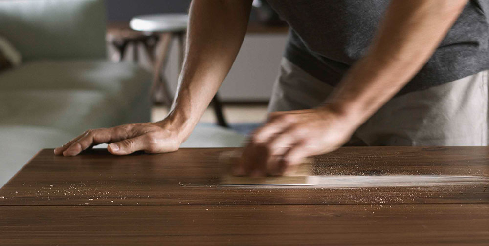 Eine Person schleift eine Naturholzoberfläche mit einem Schleifblock. Die Handbewegung erzeugt Sägespäne auf dem glänzenden Holz, was auf eine sorgfältige Pflege oder Restaurierung hinweist. Im Hintergrund ist unscharf ein Teil eines Raumes mit weiteren Möbelstücken erkennbar.