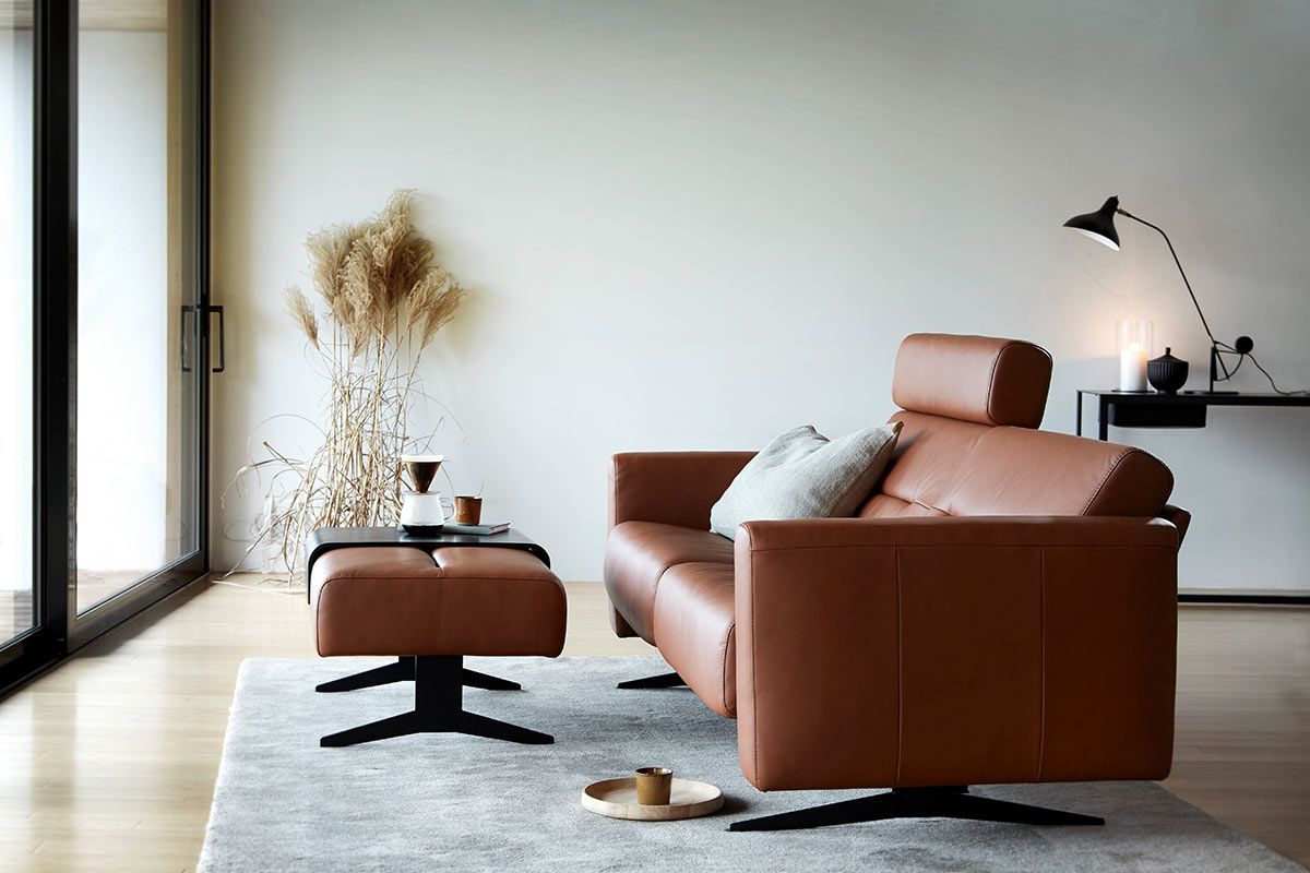 Das Bild zeigt ein modernes Wohnzimmer mit einer minimalistischen Einrichtung. Im Vordergrund steht ein braunes Ledersofa mit einem passenden Fußhocker, beide auf schwarzen Metallgestellen. Auf dem Sofa liegt ein graues Dekokissen. Vor dem Sofa steht ein kleiner runder Beistelltisch mit einer Tasse und einem Untersetzer. Rechts hinten befindet sich ein weiterer Beistelltisch mit einer brennenden Kerze und einer schwarzen Leselampe, die für zusätzliches Licht sorgt. Links im Bild sind große Glastüren zu sehen, die viel natürliches Licht hereinlassen. In der Ecke stehen getrocknete Schilfrohrgräser, die dem Raum eine natürliche Note verleihen. Der Boden ist aus hellem Holz und der Bereich um das Sofa ist mit einem hellgrauen Teppich bedeckt.