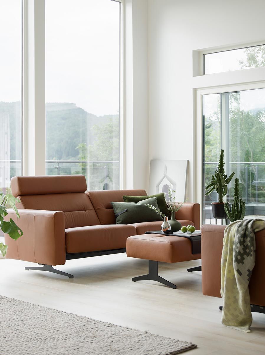 Das Bild zeigt ein modernes Wohnzimmer mit großen Fenstern, die viel natürliches Licht hereinlassen. Im Vordergrund befindet sich ein elegantes Ledersofa in einem warmen Braunton, begleitet von einem passenden Fußhocker. Auf dem Sofa liegen grüne Dekokissen, und ein kleiner Beistelltisch mit einer Blumenvase und einigen dekorativen Gegenständen steht davor. Im Hintergrund sind ein hoher Kaktus, weitere Zimmerpflanzen und ein gerahmtes Bild zu sehen, das lässig an die Wand gelehnt ist. Die helle und minimalistische Einrichtung wird durch die Aussicht ins Freie, die durch die großen Fenster sichtbar ist, ergänzt.