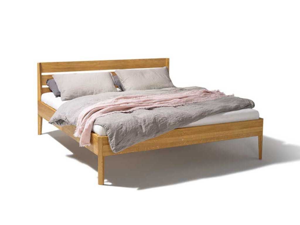 Stilvolles, minimalistisches Bett aus hellem Eichenholz mit einem schlichten, rechteckigen Kopfteil und geraden, schlanken Beinen. Das Bett ist mit neutralen, hellgrauen Bettdecken und zwei grauen Kissen bezogen, über die eine rosa Decke locker drapiert ist. Die klare Linienführung und die warme Holzoptik verleihen dem Bett ein modernes und zugleich natürliches Aussehen. Dieses Bett eignet sich ideal für ein zeitgenössisches Schlafzimmerdesign und vermittelt eine harmonische, beruhigende Atmosphäre.