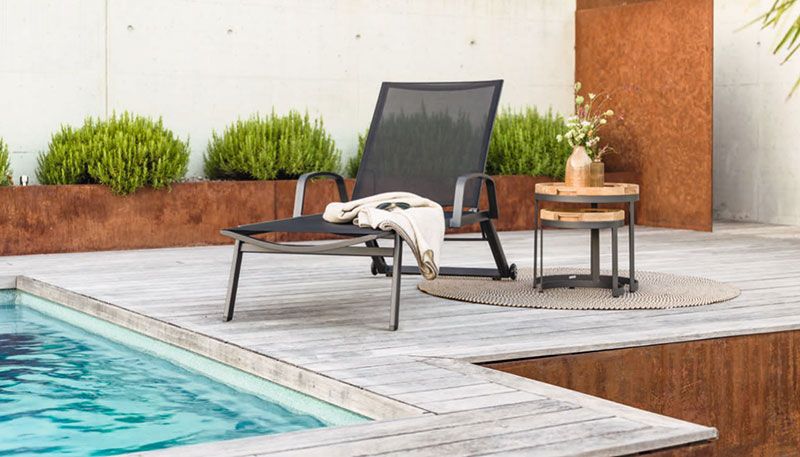 Ein moderner Liegestuhl mit schwarzer Bespannung steht an einem Pool auf einer Holzterrasse, daneben ein kleiner runder Beistelltisch aus Holz mit einem Blumenarrangement obenauf und einem gefalteten Tuch darunter. Das Ensemble ist umgeben von einer dezenten Gartenlandschaft mit sauber geschnittenen Büschen und einer rostfarbenen Wand im Hintergrund.